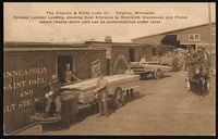 A wagon loaded with lumber sits in the middle ground, with workers preparing to load it into a one-story warehouse on the left. More forehouses are visible in the background.